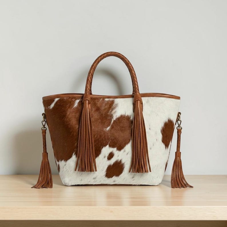 May include: A cowhide tote bag with a brown and white spotted pattern. The bag has a braided brown handle and brown leather trim. Brown leather tassels hang from the sides. The bag sits on a light brown wooden surface.
