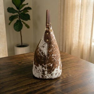 May include: A cowhide shoulder bag with a brown leather handle and accents. The bag features a spotted pattern of brown and white. The bag is sitting on a dark wooden surface.