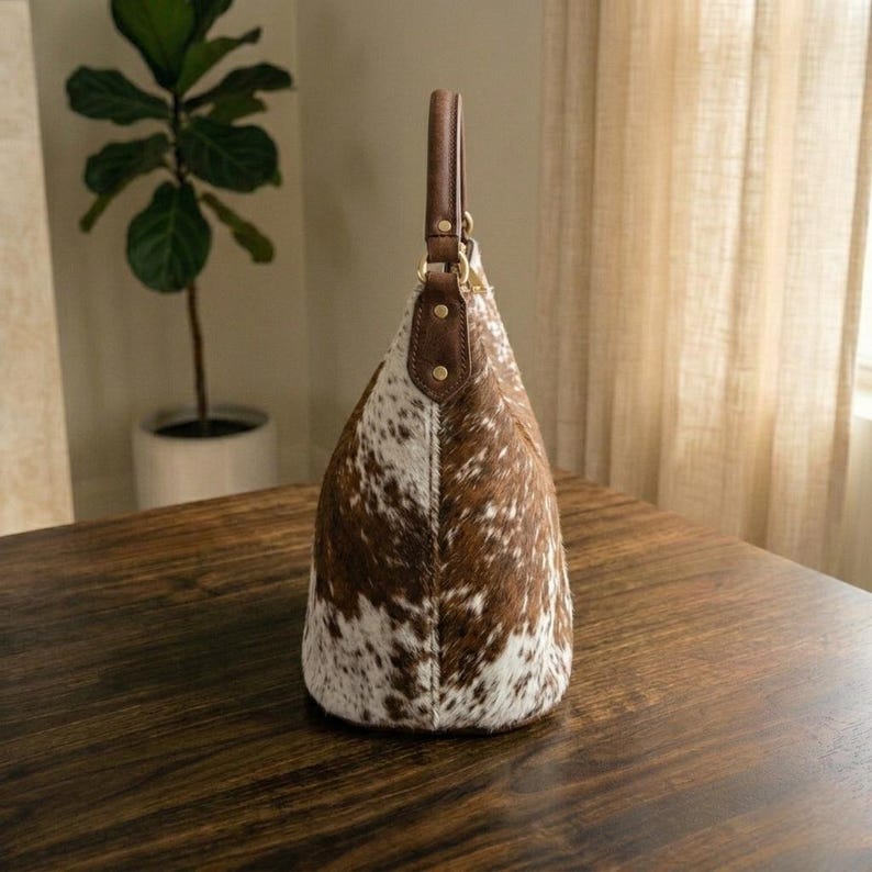 May include: A cowhide shoulder bag with a brown leather handle and accents. The bag features a spotted pattern of brown and white. The bag is sitting on a dark wooden surface.