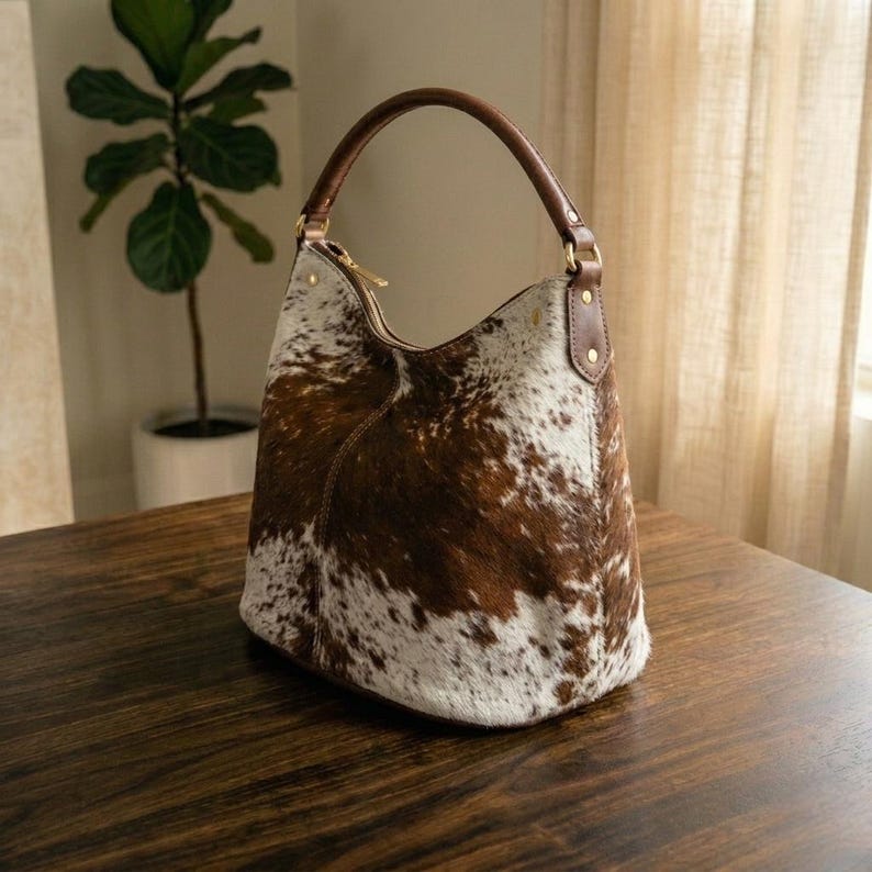 May include: A brown and white cowhide leather hobo bag with a brown leather handle and gold zipper. The bag is on a dark wooden surface. The bag has a rounded shape and a single handle.