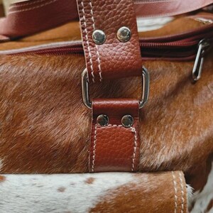 Puede incluir: Un bolso de viaje de piel de vaca marr&oacute;n y blanco con una correa de cuero texturizado sujeta con remaches met&aacute;licos plateados y una anilla rectangular. El bolso incluye un cierre de cremallera y un bolsillo frontal con pelaje moteado.