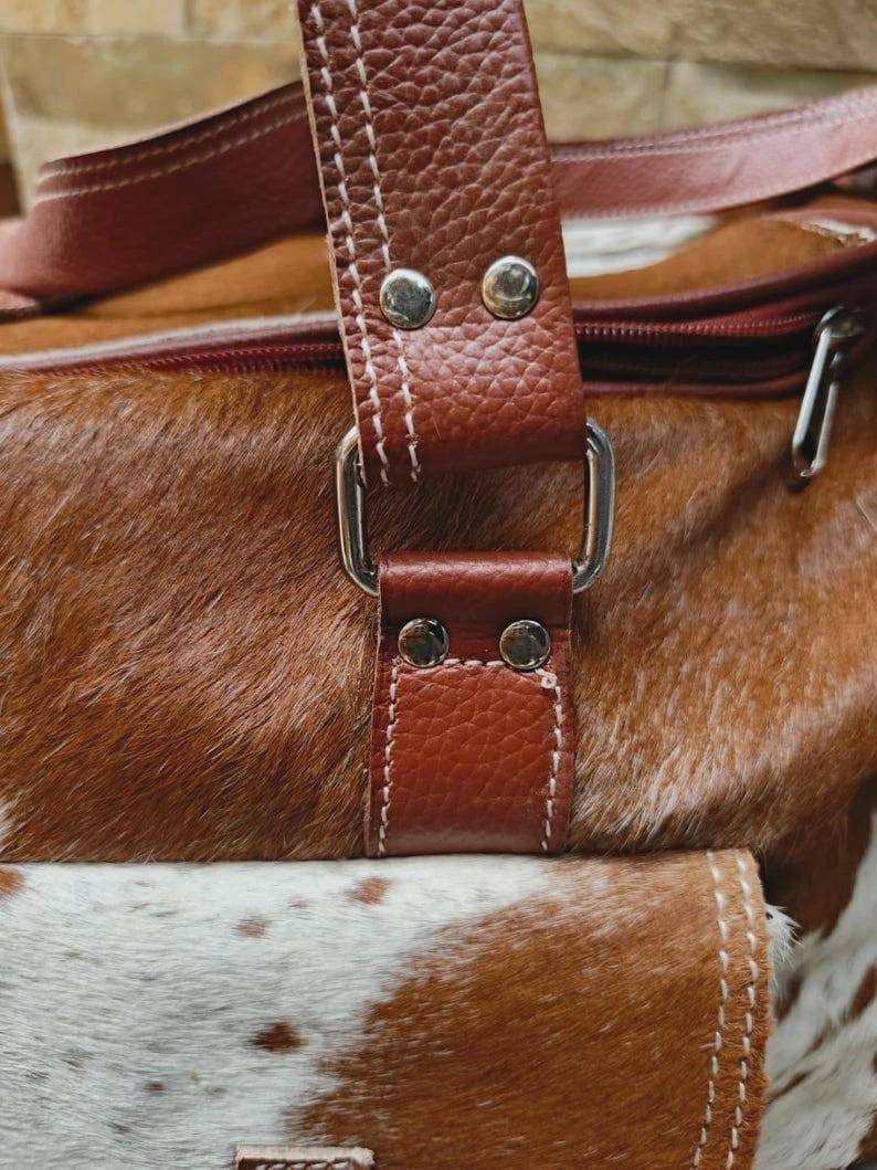 Puede incluir: Un bolso de viaje de piel de vaca marr&oacute;n y blanco con una correa de cuero texturizado sujeta con remaches met&aacute;licos plateados y una anilla rectangular. El bolso incluye un cierre de cremallera y un bolsillo frontal con pelaje moteado.