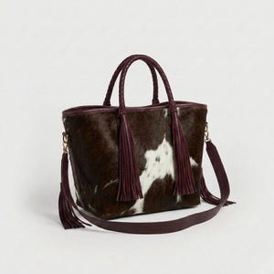 May include: A brown and white cowhide tote bag with braided handles and a detachable shoulder strap. The bag features burgundy leather trim and tassels, with gold-coloured hardware. The bag is designed for carrying items.