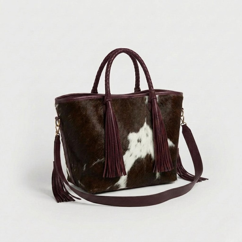 May include: A brown and white cowhide tote bag with braided handles and a detachable shoulder strap. The bag features burgundy leather trim and tassels, with gold-coloured hardware. The bag is designed for carrying items.