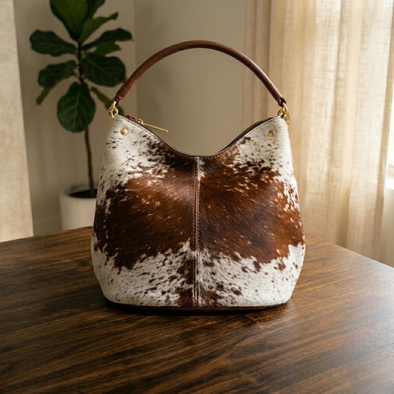 May include: A cowhide leather handbag with a brown handle and gold-colored hardware. The bag features a unique pattern of brown and white patches. The bag is sitting on a wooden surface.