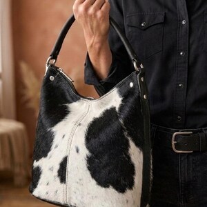 K&ouml;nnte beinhalten: Eine Schultertasche im Hobo-Stil mit schwarz-wei&szlig;em Kuhfellmuster und einer texturierten Oberfl&auml;che aus echtem Fell. Die Tasche verf&uuml;gt &uuml;ber einen einzelnen schwarzen Ledergriff mit silberfarbenen Metallbeschl&auml;gen, einen Rei&szlig;verschluss und sichtbare N&auml;hte.