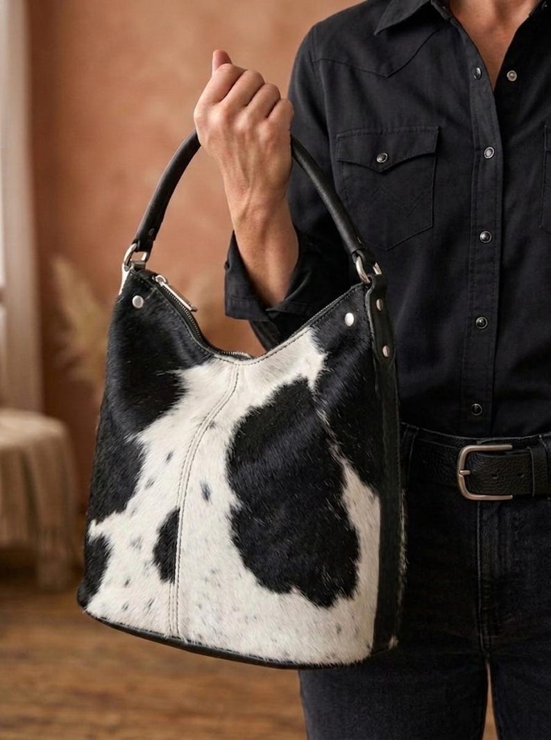 K&ouml;nnte beinhalten: Eine Schultertasche im Hobo-Stil mit schwarz-wei&szlig;em Kuhfellmuster und einer texturierten Oberfl&auml;che aus echtem Fell. Die Tasche verf&uuml;gt &uuml;ber einen einzelnen schwarzen Ledergriff mit silberfarbenen Metallbeschl&auml;gen, einen Rei&szlig;verschluss und sichtbare N&auml;hte.