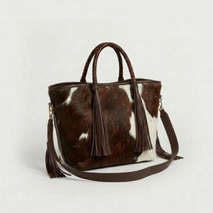 May include: A brown and white cowhide tote bag with braided handles and a detachable shoulder strap. The bag features two long brown tassels and gold-coloured hardware. The bag is designed with a rectangular shape.