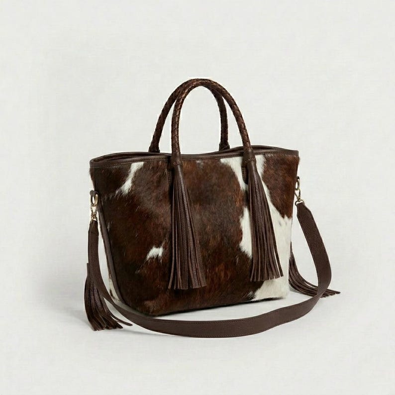 May include: A brown and white cowhide tote bag with braided handles and a detachable shoulder strap. The bag features two long brown tassels and gold-coloured hardware. The bag is designed with a rectangular shape.