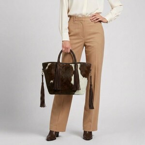May include: A brown and white cowhide tote bag with a braided handle and long brown tassels. The bag is held by a person wearing a white blouse and tan trousers. The bag has a shoulder strap.