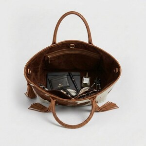May include: Open tote bag with brown leather handles and a cowhide pattern. The interior is brown suede and contains a wallet, pen, notebook, perfume, keys, and a scarf. Tassels hang from the sides.