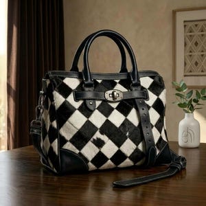 May include: A handbag featuring a black and white checkered pattern made of textured hair-on hide. The bag includes black leather trim, two top handles, a central metal turn-lock closure on a horizontal strap, and a detachable shoulder strap.
