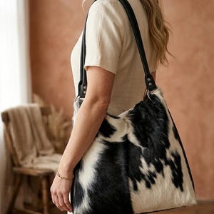 May include: A large, black and white cowhide shoulder bag with a black leather strap. The bag has a soft, textured appearance and a large capacity. The strap is attached to the bag with silver metal rings.