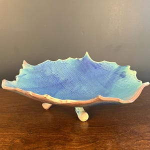 May include: A ceramic bowl shaped like a leaf with a textured surface, featuring a vibrant blue and turquoise glaze. The bowl has a scalloped edge with a light brown rim and is supported by three small, matching ceramic feet on the underside.