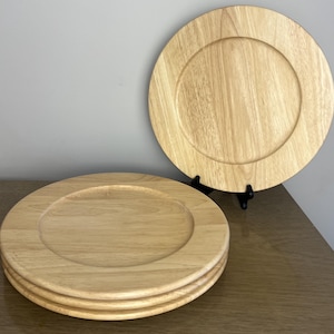 Peut inclure: Un ensemble de trois assiettes rondes en bois avec une finition naturelle en grain de bois brun clair. Deux assiettes sont empilées horizontalement, tandis que la troisième est placée verticalement derrière, soutenue par un support en plastique noir.