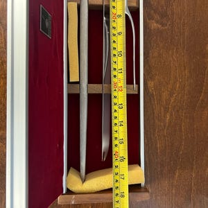 May include: A silver-toned metal carving fork and knife set housed in a rectangular wooden case with a deep red velvet lining. A yellow measuring tape is placed over the utensils, indicating a length of approximately 18 inches for the set.