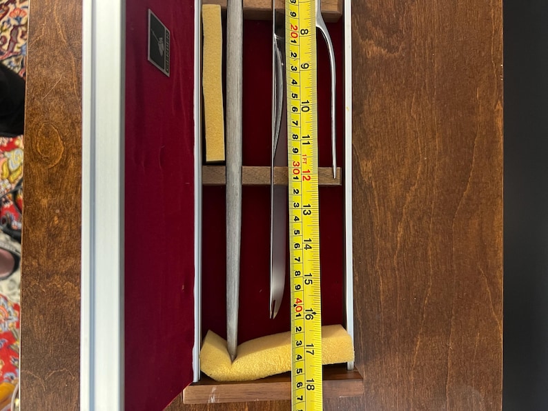 May include: A silver-toned metal carving fork and knife set housed in a rectangular wooden case with a deep red velvet lining. A yellow measuring tape is placed over the utensils, indicating a length of approximately 18 inches for the set.