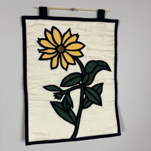 Peut inclure: Une tenture murale rectangulaire en tissu représentant une grande fleur jaune avec un centre sombre et des feuilles vertes sur un fond crème. La pièce est encadrée par une bordure noire épaisse et suspendue à une tige en bois par deux boucles.