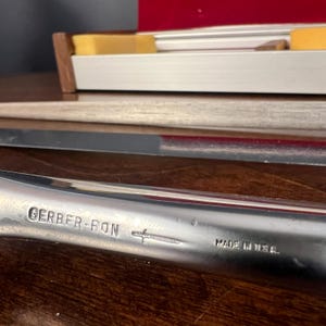 May include: A silver-toned metal knife blade with a polished finish. The handle area is engraved with the text GERBER-RON, a small sword icon, and the words MADE IN U.S.A. A partially visible storage box with yellow foam inserts is in the background.