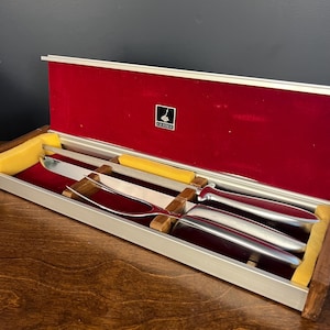May include: A silver-toned carving knife and a two-pronged carving fork with smooth metal handles, presented in a rectangular case with a red velvet interior, yellow foam inserts, and a small black and white Gerber logo plaque on the lid.
