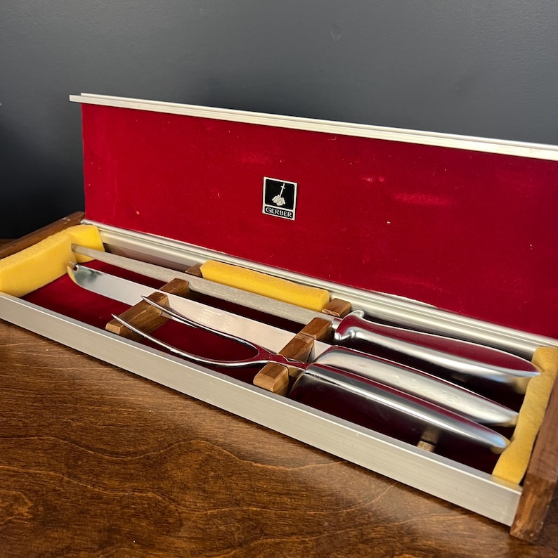 May include: A silver-toned carving knife and a two-pronged carving fork with smooth metal handles, presented in a rectangular case with a red velvet interior, yellow foam inserts, and a small black and white Gerber logo plaque on the lid.