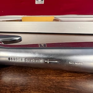 May include: A silver-colored metal blade featuring the engraved text GERBER GUNGNIR with a small sword icon and the text MADE IN USA. The metallic surface has a smooth finish and is positioned horizontally, showing clear branding and manufacturing details.