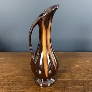 May include: A tall, slender ceramic pitcher vase featuring a glossy dark brown glaze with vertical streaks of light tan or cream color. The vessel has a rounded base, a long, curved neck, and a single handle that extends from the rim to the body.