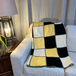 May include: A handmade, square-patterned blanket in black, white, and yellow, draped over a white sofa. The blanket features a checkerboard design with textured squares and a green border. The blanket is a cosy home decor item.