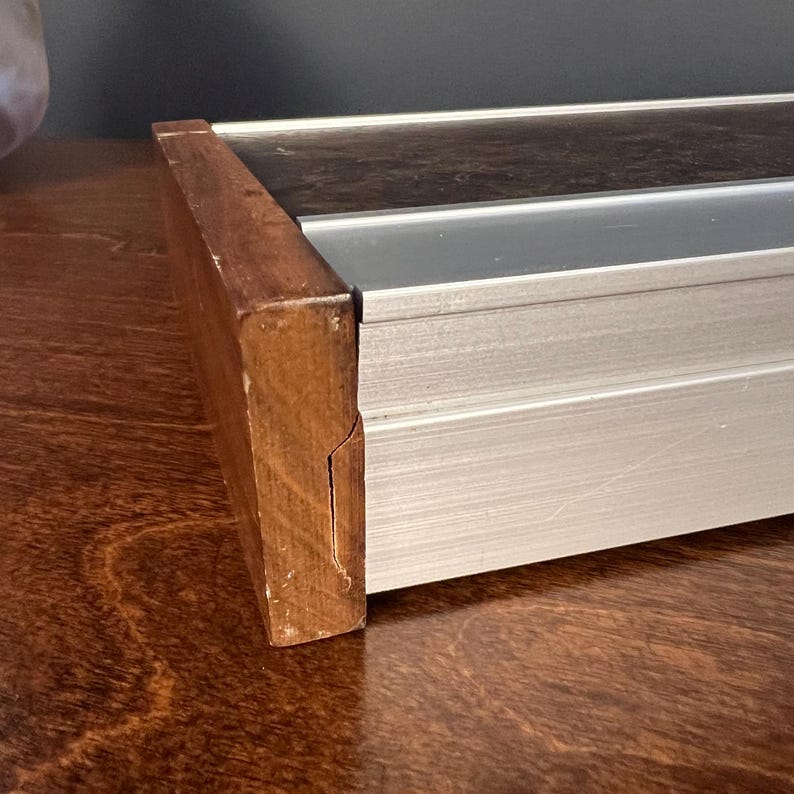 May include: A rectangular object featuring a brushed silver aluminum frame with a dark brown, textured top surface. The side of the object is finished with a solid brown wooden end cap that shows a visible vertical crack along its side surface.