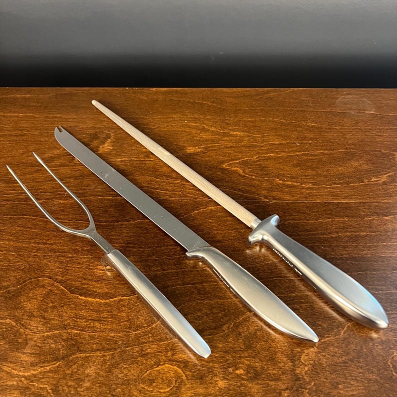 May include: A three-piece carving set consisting of a two-pronged carving fork, a long straight-edged carving knife, and a sharpening steel rod. Each tool features a smooth, polished silver-tone metal handle with a sleek, tapered design.