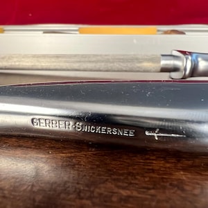 May include: A silver-toned metal carving knife with a smooth, polished handle. The blade features the engraved text GERBER SNICKERSNEE followed by a small sword icon. The knife is positioned horizontally with a sharpening steel visible behind it.