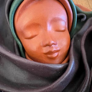 Könnte beinhalten: Eine terrakottafarbene Skulptur eines ruhigen Gesichts mit geschlossenen Augen, umrahmt von einem dunkelvioletten, grünen und orangefarbenen Stoff. Die Skulptur hat glatte Züge und einen friedlichen Ausdruck, der auf ein religiöses oder spirituelles Thema hindeutet.