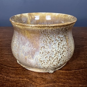 May include: A round ceramic bowl with a wide, flared rim. The exterior features a mottled glaze with blended tones of earthy brown, muted blue, and cream, creating a textured, speckled appearance across the curved surface of the vessel.