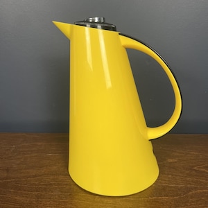 Puede incluir: Una jarra térmica de color amarillo brillante con un cuerpo cónico, un asa negra y una tapa de metal plateado. El recipiente cuenta con un pico vertedor puntiagudo y un acabado exterior liso de color sólido, diseñado para contener líquidos.