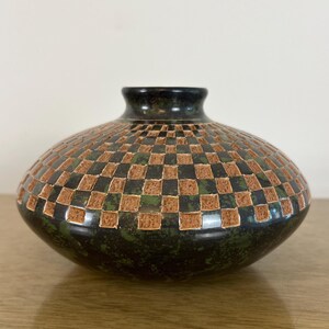 Puede incluir: Un jarrón de cerámica bajo y redondeado con un esmalte moteado en verde oscuro y negro. La parte superior del jarrón está decorada con un patrón de tablero de ajedrez texturizado y repetitivo que alterna entre cuadrados oscuros lisos y cuadrados beige hundidos.