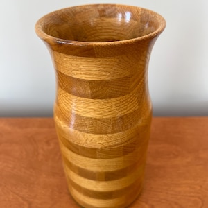 Könnte beinhalten: Eine Holzvase mit einem hellbraunen und mittelbraunen Streifenmuster. Die Vase hat einen ausgestellten oberen Rand und einen zylindrischen Körper. Die Holzmaserung ist sichtbar und verleiht dem Stück Textur. Die Vase ist wahrscheinlich für dekorative Zwecke.