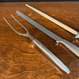 May include: A stainless steel carving fork with a long, two-pronged head and a smooth, solid handle, positioned alongside a long, thin carving knife with a matching handle and a sharpening steel featuring a light-colored wooden handle.