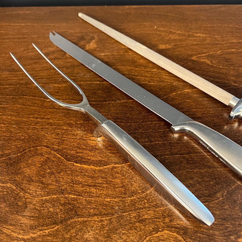 May include: A stainless steel carving fork with a long, two-pronged head and a smooth, solid handle, positioned alongside a long, thin carving knife with a matching handle and a sharpening steel featuring a light-colored wooden handle.