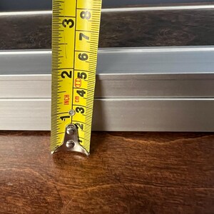 May include: A silver-colored metal extrusion profile with a rectangular cross-section. A yellow measuring tape is positioned vertically against the object, showing a height measurement of approximately 3.5 inches for the metal piece.