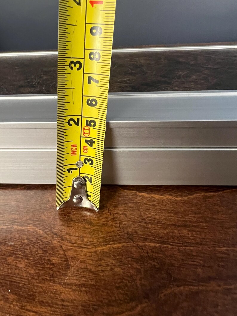 May include: A silver-colored metal extrusion profile with a rectangular cross-section. A yellow measuring tape is positioned vertically against the object, showing a height measurement of approximately 3.5 inches for the metal piece.