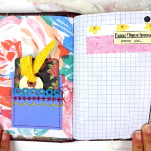 May include: An open journal with a blue pocket on the left page. The pocket has a yellow ribbon and a tag with a floral design. The right page has a pink and white floral patterned sticker with the text "Eldridge E. Wheeler Seedsman, Bridgeport, Conn."
