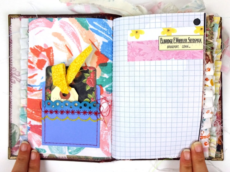 May include: An open journal with a blue pocket on the left page. The pocket has a yellow ribbon and a tag with a floral design. The right page has a pink and white floral patterned sticker with the text "Eldridge E. Wheeler Seedsman, Bridgeport, Conn."