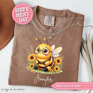 May include: A brown t-shirt featuring a graphic of a yellow bee wearing black-rimmed glasses, surrounded by sunflowers. The name Jennifer is printed in white script below the bee. The shirt has a Comfort Colors label at the inner neckline.