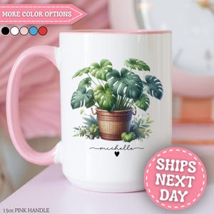 May include: A white ceramic mug with a pink handle and interior rim, featuring a graphic of a green monstera plant in a brown woven basket. The name michelle is printed in black script below the plant, accompanied by a small black heart icon.