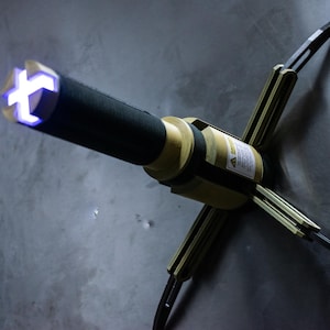 Op de afbeelding: Een futuristisch apparaat met een zwarte cilindrische behuizing en een blauw, kruisvormig licht aan de punt. Het apparaat is gemonteerd op een metalen oppervlak met vier uitklapbare armen. De behuizing is beige en zwart.