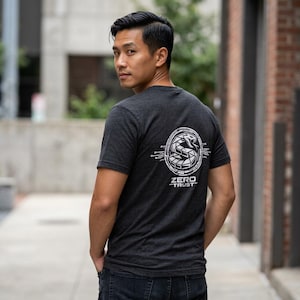 May include: A charcoal gray short-sleeve t-shirt featuring a white graphic on the back. The design displays a stylized snake coiled within a circular circuit board pattern, with the text ZERO TRUST printed in white capital letters underneath.