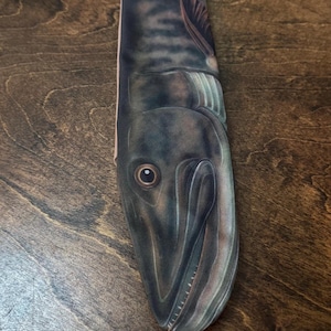 May include: A long, narrow sock featuring a detailed illustration of a fish. The fish is primarily dark gray and brown with a detailed eye and fin. The sock is displayed on a wooden surface.