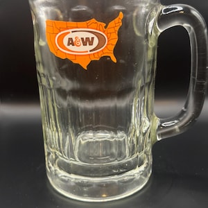 May include: Clear glass beer mug with a handle and a rectangular shape. The mug features the A&W logo in a brown and orange design, with the outline of the United States. The mug is empty and sits on a dark surface.