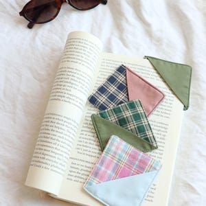 Puede incluir: Un libro abierto con varios marcadores de esquina de tela a cuadros de colores. Los marcadores son en tonos de azul, rosa, verde y verde oliva. Gafas de sol carey descansan sobre el fondo de tela blanca.