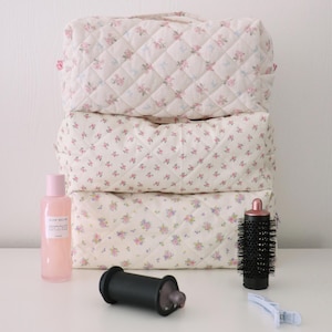 May include: A stack of three quilted fabric pouches featuring a cream background with a small pink and blue floral pattern. Beside the pouches are a pink bottle of Glow Recipe toner, a black cylindrical hair roller, a black round brush, and a light blue clip.